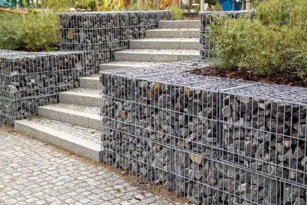 Gabion Retaining Walls by Retaining Walls Tauranga, used to frame garden beds and support stairs with stacked wire cages filled with dark stones.